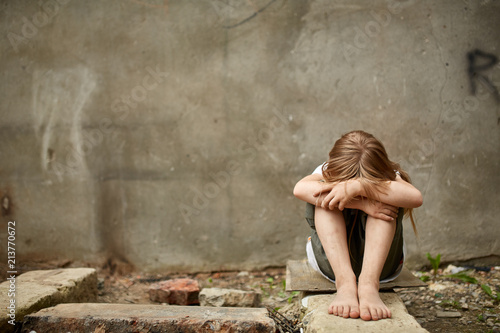 Street photo of girl orphan with holen knees under the dirty city wall.
