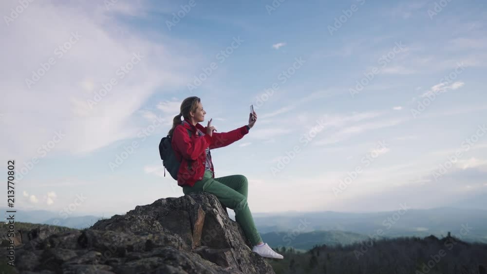 the traveler takes a selfie in the mountains. young female tourist sitting on the edge of a cliff and taking pictures on the camera smartphone the traveler takes a selfie in the mountains. young female tourist sitting on the edge of a cliff and taking pictures on the camera smartphone