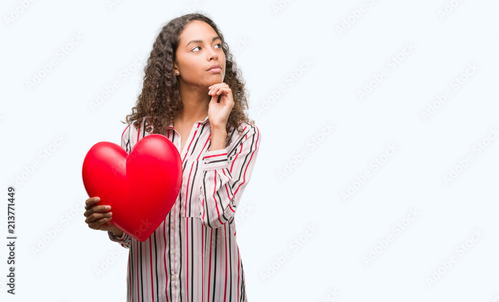Young hispanic woman in love holding red heart serious face thinking about question, very confused idea Young hispanic woman in love holding red heart serious face thinking about question, very confused idea