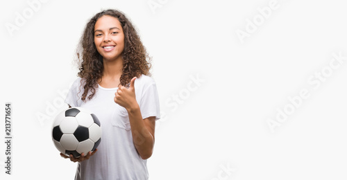 Young hispanic woman holding soccer football ball happy with big smile doing ok sign, thumb up with fingers, excellent sign