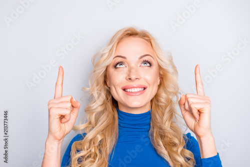 Portrait of stylish, pretty, trendy,charming, nice, sexy, cute, blonde woman in blue roll neck pointing two forefingers up isolated on grey background looking at advertisement, copy space, empty place