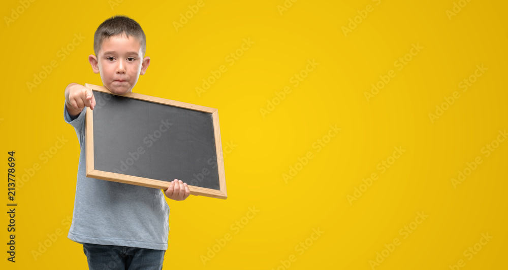 Dark haired little child holding a blackboard pointing with finger to the camera and to you, hand sign, positive and confident gesture from the front Dark haired little child holding a blackboard pointing with finger to the camera and to you, hand sign, positive and confident gesture from the front