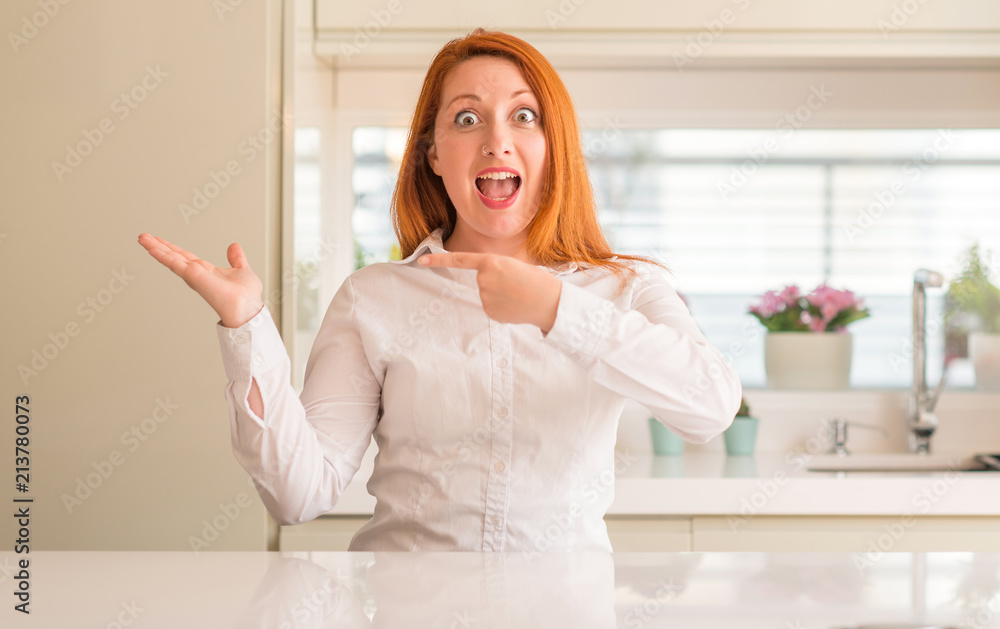 Redhead woman at kitchen amazed and smiling to the camera while presenting with hand and pointing with finger. Redhead woman at kitchen amazed and smiling to the camera while presenting with hand and pointing with finger.