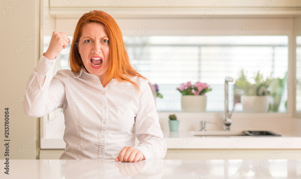Redhead woman at kitchen angry and mad raising fist frustrated and furious while shouting with anger. Rage and aggressive concept.