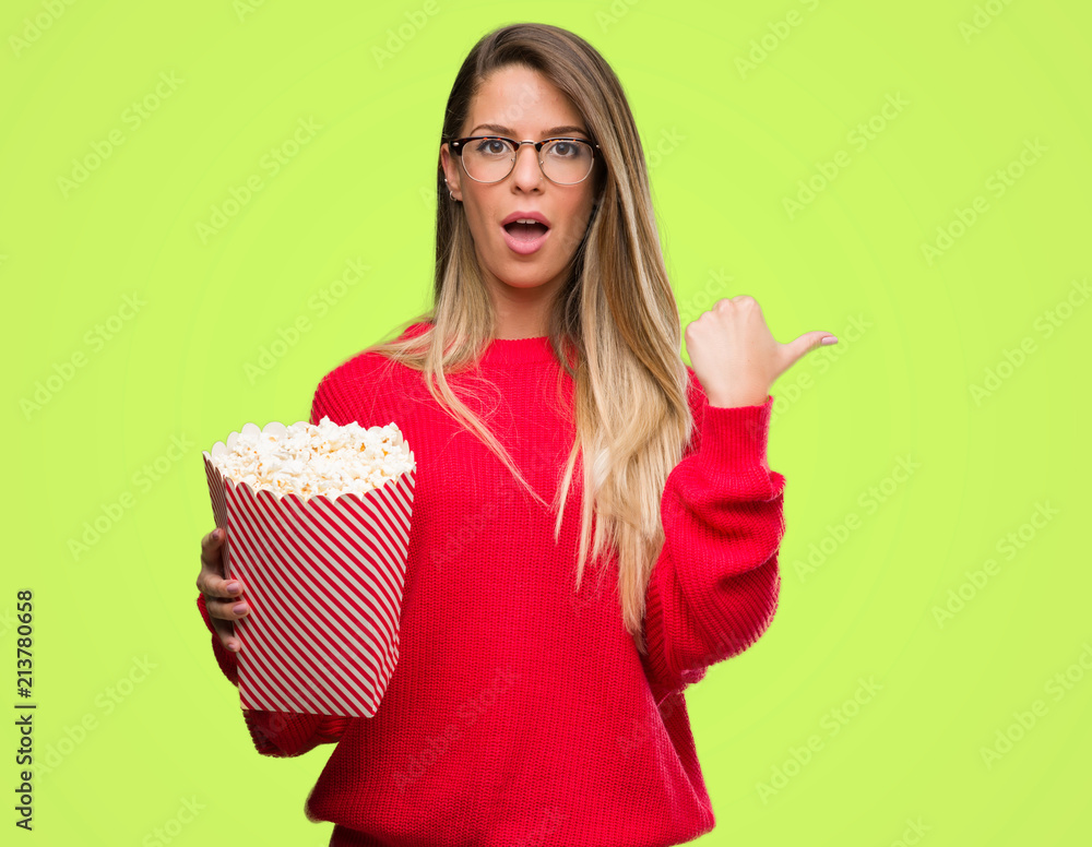 Beautiful young woman eating popcorn pointing with hand and finger up with happy face smiling Beautiful young woman eating popcorn pointing with hand and finger up with happy face smiling