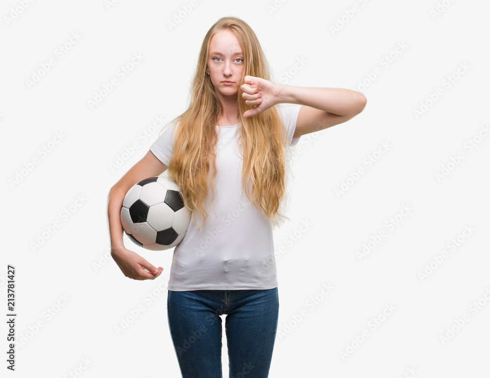 Blonde teenager woman holding soccer football ball with angry face, negative sign showing dislike with thumbs down, rejection concept