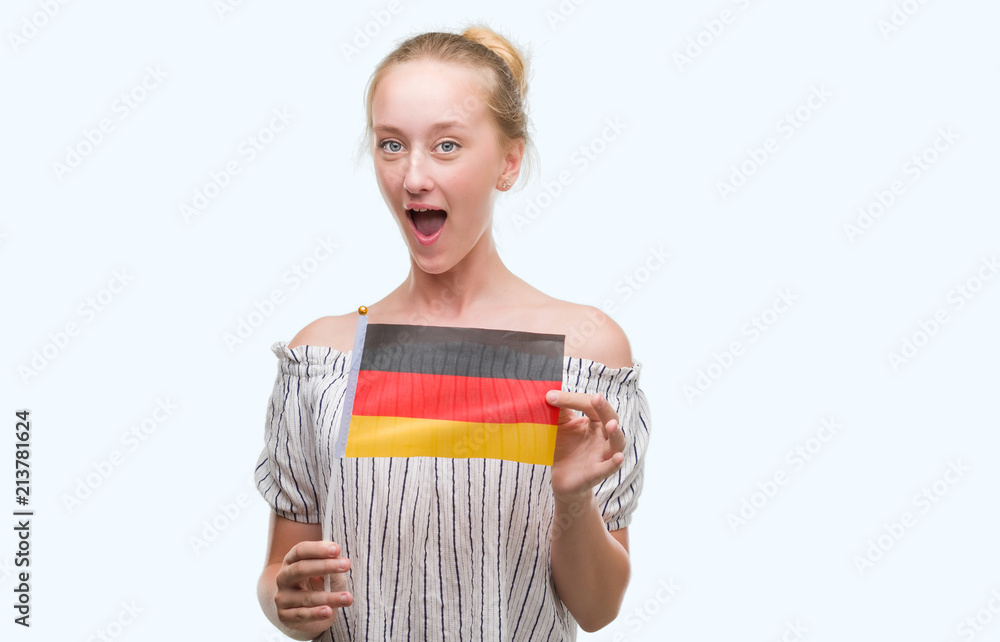 Blonde teenager woman holding flag of Germany scared in shock with a surprise face, afraid and excited with fear expression Blonde teenager woman holding flag of Germany scared in shock with a surprise face, afraid and excited with fear expression