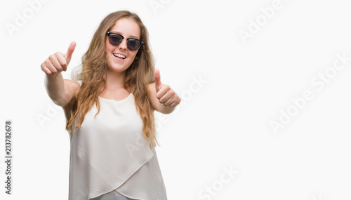 Young blonde woman wearing sunglasses approving doing positive gesture with hand, thumbs up smiling and happy for success. Looking at the camera, winner gesture. © Krakenimages.com