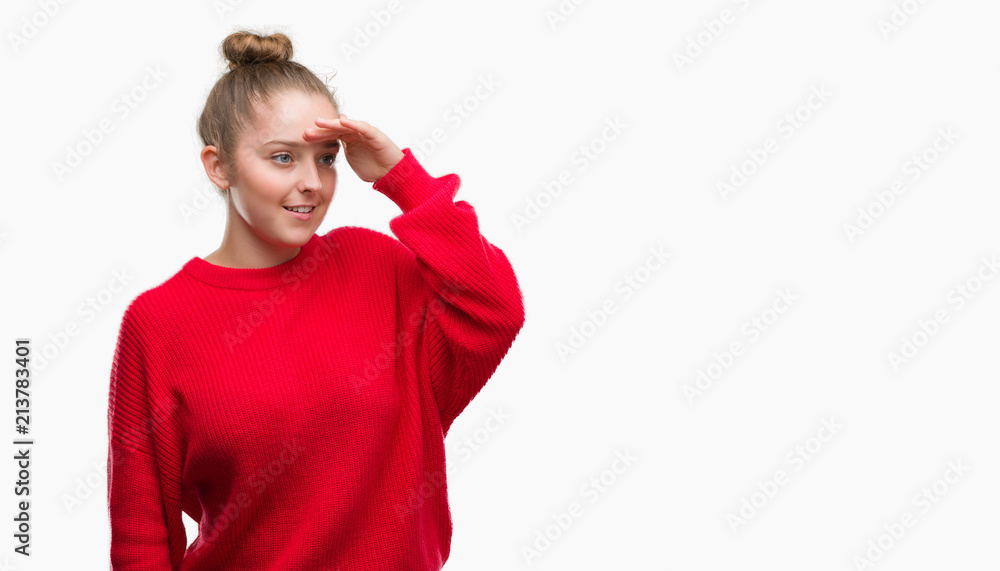 Young blonde woman wearing bun and red sweater very happy and smiling looking far away with hand over head. Searching concept. Young blonde woman wearing bun and red sweater very happy and smiling looking far away with hand over head. Searching concept.