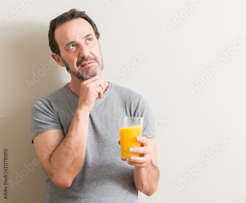 Senior man drinking orange juice in a glass serious face thinking about question, very confused idea