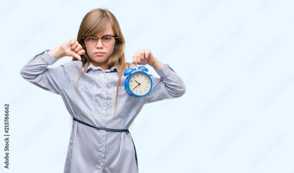 Fototapeta premium Young blonde child holding alarm clock with angry face, negative sign showing dislike with thumbs down, rejection concept