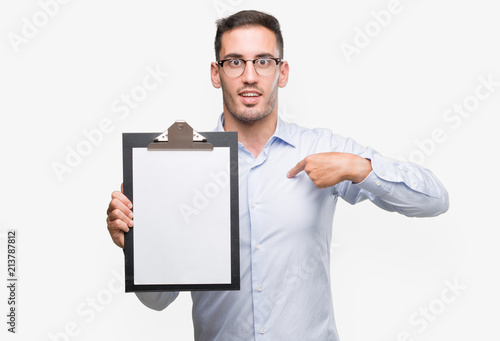 Handsome young business man showing a clipboard with surprise face pointing finger to himself