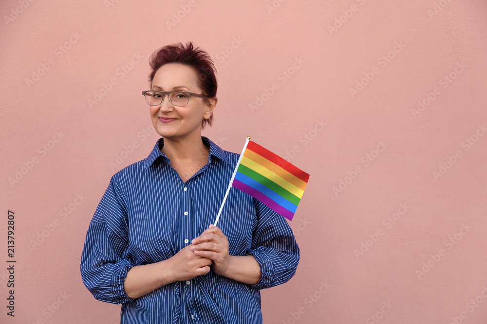 LGBT flag. Woman holding rainbow flag. Nice portrait of middle aged lady 40 50 years old with a flag over pink wall background. LGBT flag. Woman holding rainbow flag. Nice portrait of middle aged lady 40 50 years old with a flag over pink wall background.