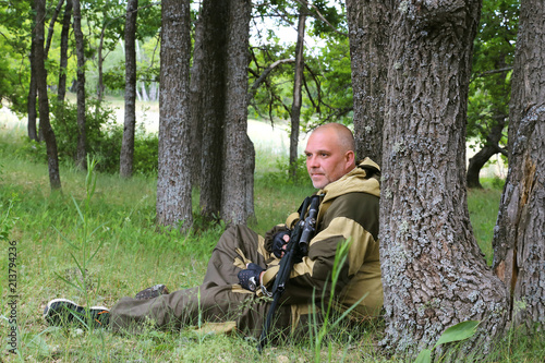 A hunter man with a firearm sits in a forest under a tree. Hunting season.