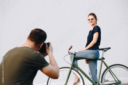 Professional photographer doing a photo shooting of young stylish girl sitting on bike and posing at camera, against the white wall background. Outdoors.