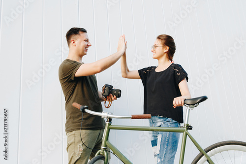 Profile photo of cheerful guy with camera and gorgeous girl with bike, giving a high five each other, after photo shooting outdoors.