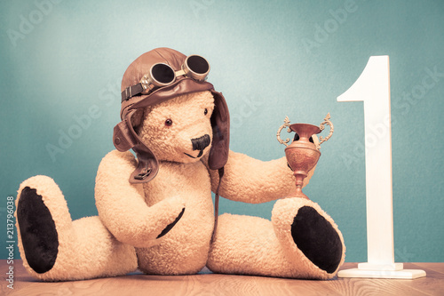 Retro Teddy Bear toy in leather pilot's hat and vintage goggles sitting with award winner's trophy cup and big wooden number one front mint green wall background. Old style filtered photo