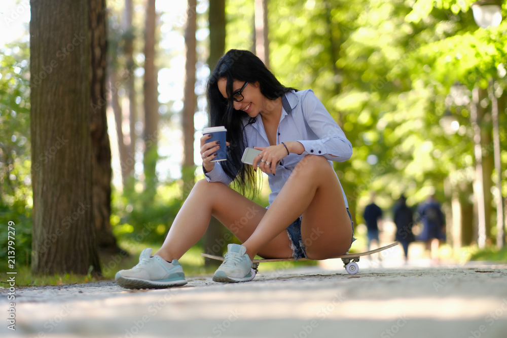 Naklejka premium Laughing sexy brunette girl in glasses wearing a shirt and shorts sitting on a skateboard and holds a cup of takeaway coffee and smartphone at the park.