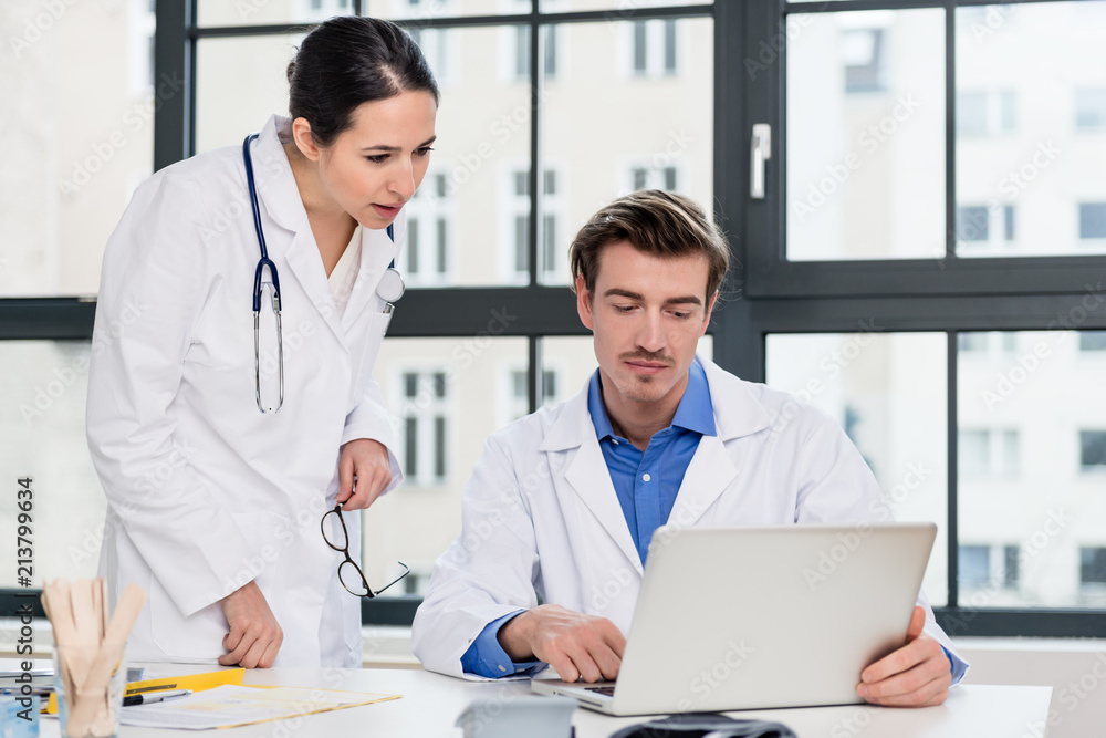 Young female physician using a tablet PC while asking for advice from her experienced male colleague in the office a modern hospital Young female physician using a tablet PC while asking for advice from her experienced male colleague in the office a modern hospital