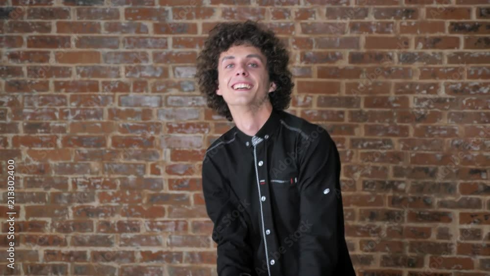 Young handsome cheerful man with curly hair dancing and looking at camera, brick wall background Young handsome cheerful man with curly hair dancing and looking at camera, brick wall background