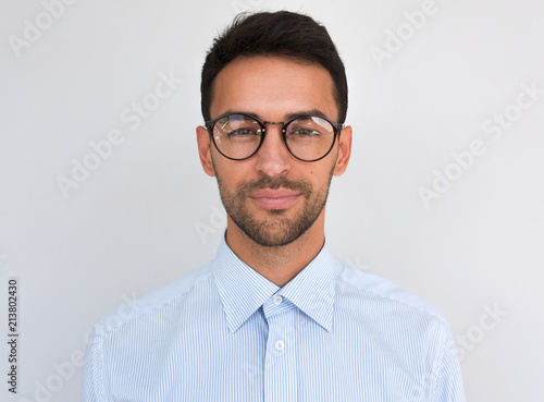 Closeup headshot of handsome attractive male, looks directly at the camera, wears round spectacles, isolated over white background. Portrait of smart bristle student wearing casual blue shirt. People