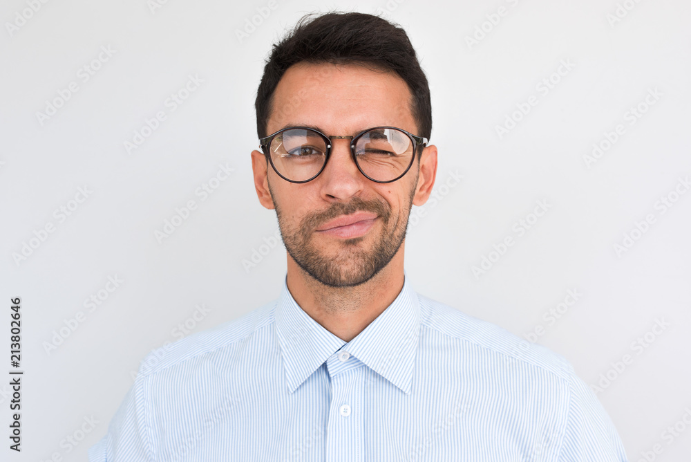 Portrait of unshaven handsome male frowns, being dissatisfied with something, blink with eye, wears round glasses and blue shirt, isolated over white studio background. People and emotion concept. Portrait of unshaven handsome male frowns, being dissatisfied with something, blink with eye, wears round glasses and blue shirt, isolated over white studio background. People and emotion concept.