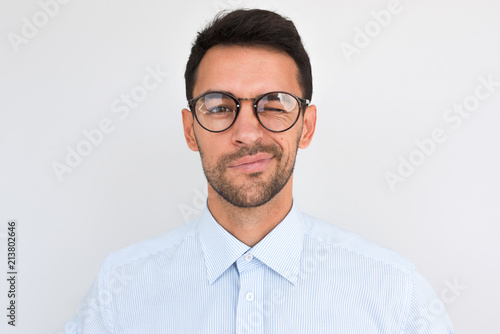 Portrait of unshaven handsome male frowns, being dissatisfied with something, blink with eye, wears round glasses and blue shirt, isolated over white studio background. People and emotion concept.