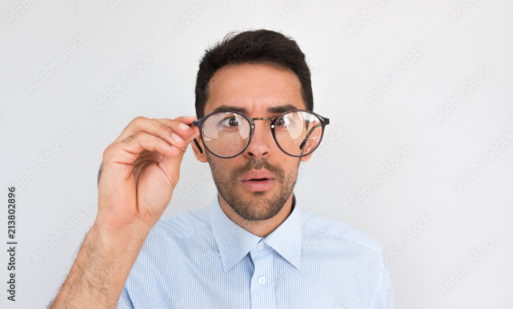 Closeup image of astonished young European handsome male, looking through spectacles, touches rim of glasses, wears blue shirt posing on white studio wall. People, emotion and lifestyle concept Closeup image of astonished young European handsome male, looking through spectacles, touches rim of glasses, wears blue shirt posing on white studio wall. People, emotion and lifestyle concept