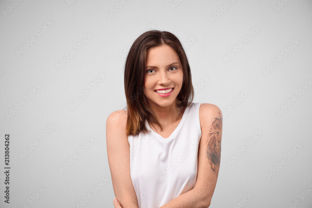 Portrait of beautiful young brunette woman with tattoo in white t-shirt looking at camera and smiling isolated on grey Studio background. Portrait of beautiful young brunette woman with tattoo in white t-shirt looking at camera and smiling isolated on grey Studio background.