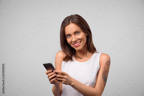 Brunette smiling young woman demonstrating white teeth using cell phone, messaging, being happy to text with her boyfriend. Modern technologies and communication
