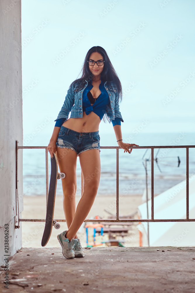 Seductive hot brunette girl wearing short shorts and jeans jacket holds a  skateboard while standing to lean on a guardrail against the sea coast.  Stock Photo | Adobe Stock
