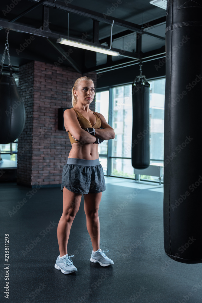 Punching bags. Blonde-haired athlete female boxer standing in gym near punching bags