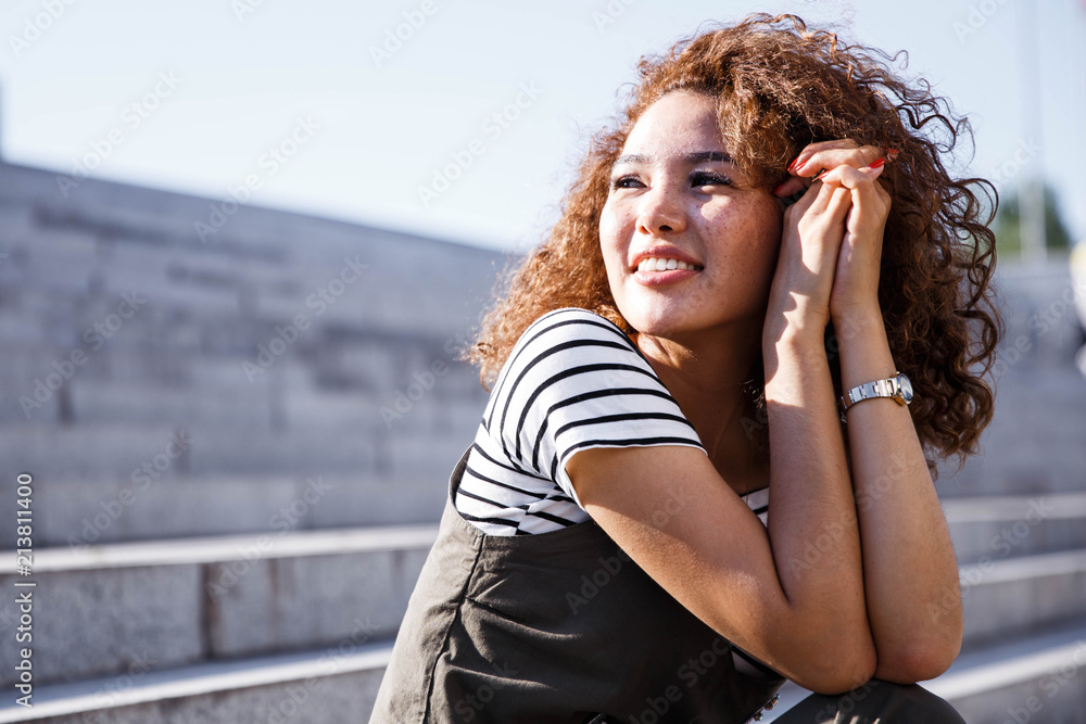 Outdoor close up portrait of young beautiful happy smiling redhead girl with freckles, long curly hair, no makeup. Model posing in street. Summer sunny day. Female natural beauty, happiness concept