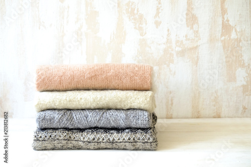 Bunch of knitted warm pastel color sweaters with different knitting patterns folded in stack on white wooden table, textured wall background. Fall winter season knitwear. Close up, copy space for text
