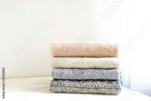 Bunch of knitted warm pastel color sweaters with different knitting patterns folded in stack on white wooden table, textured wall background. Fall winter season knitwear. Close up, copy space for text