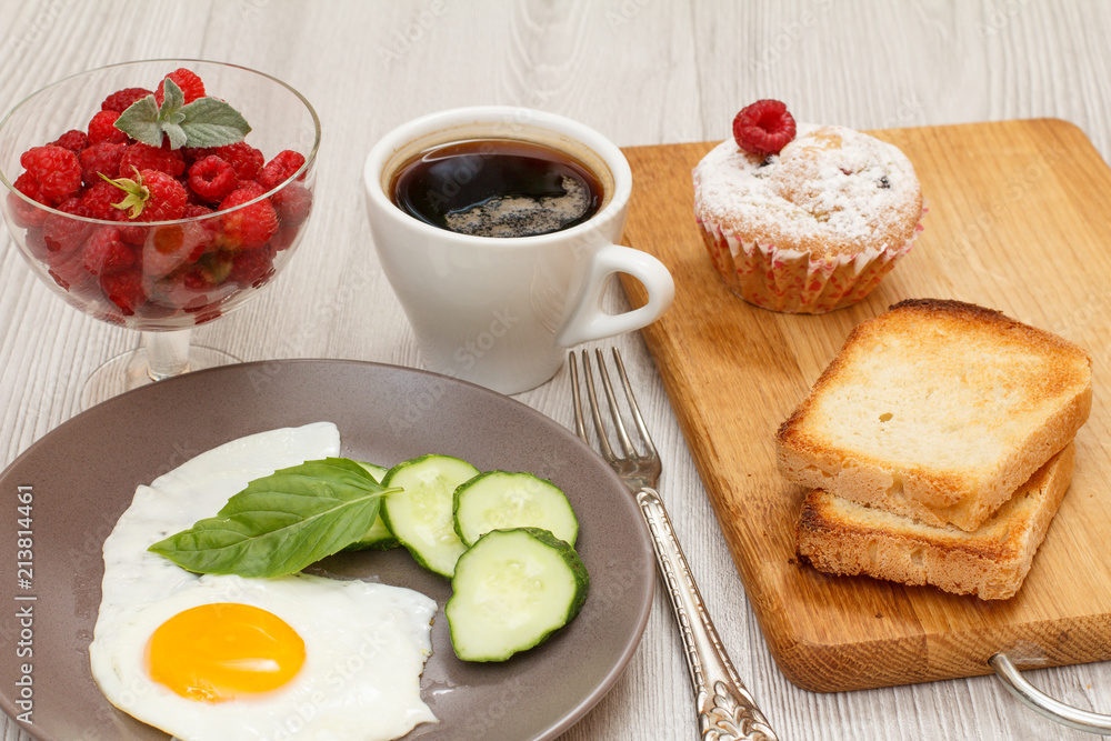 Plate with fried egg, cut cucumber and leaf of basil, glass bowl with raspberries, cup of coffee, fork, muffin and toasts on wooden cutting board. Plate with fried egg, cut cucumber and leaf of basil, glass bowl with raspberries, cup of coffee, fork, muffin and toasts on wooden cutting board.