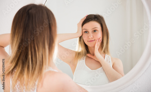 Young woman using mirror and touching face in bedroom