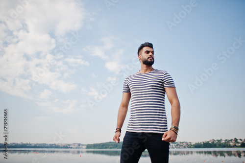 Handsome tall arabian beard man model at stripped shirt posed outdoor against lake and sky. Fashionable arab guy.