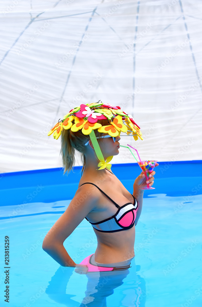 A young girl wearing a bikini and a butterfly hat holds  an imaginative cocktail drink in her hand. She is stands  side on to the camera in a swimming pool.