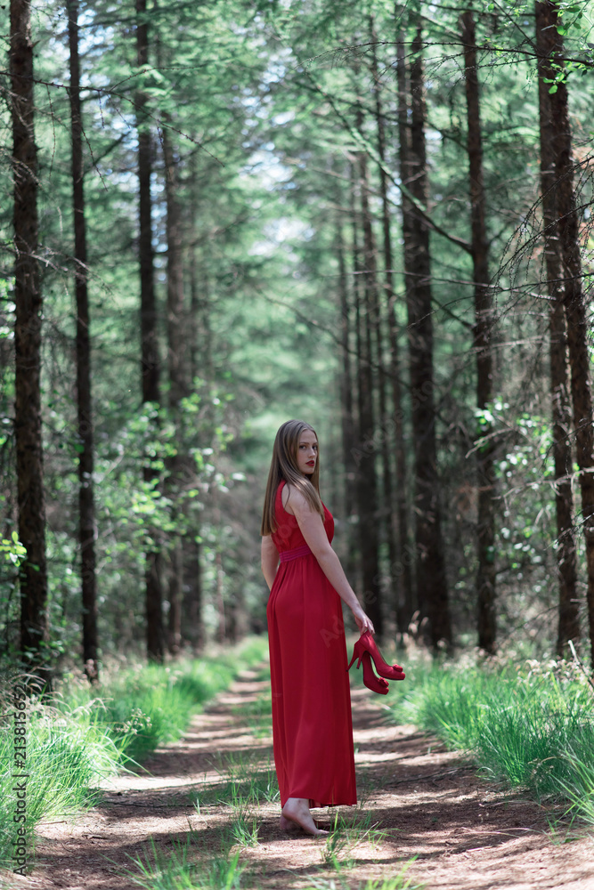 Blonde woman in red dress holding shoes in hand looking over shoulder. Walking on forest path. Blonde woman in red dress holding shoes in hand looking over shoulder. Walking on forest path.