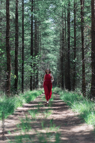Billede på lærred Blonde woman in red dress running towards camera on forest path.
