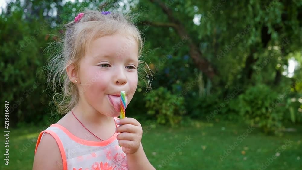 Little child girl eat lick tongue a sweet colored caramel candy stick in green garden Little child girl eat lick tongue a sweet colored caramel candy stick in green garden