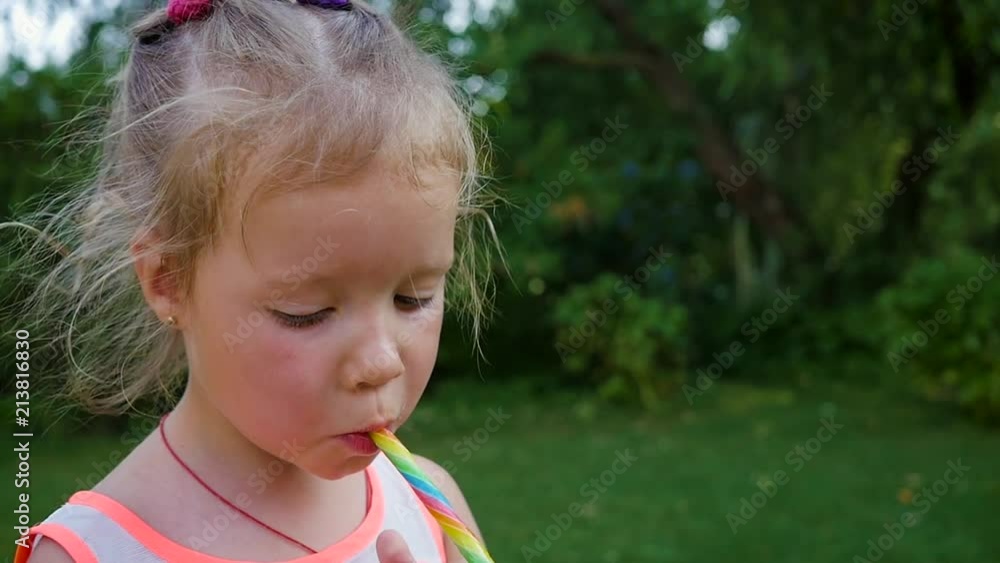 Little child girl eat lick suck a sweet colored caramel candy stick in green garden Stock ビデオ | Adobe Stock