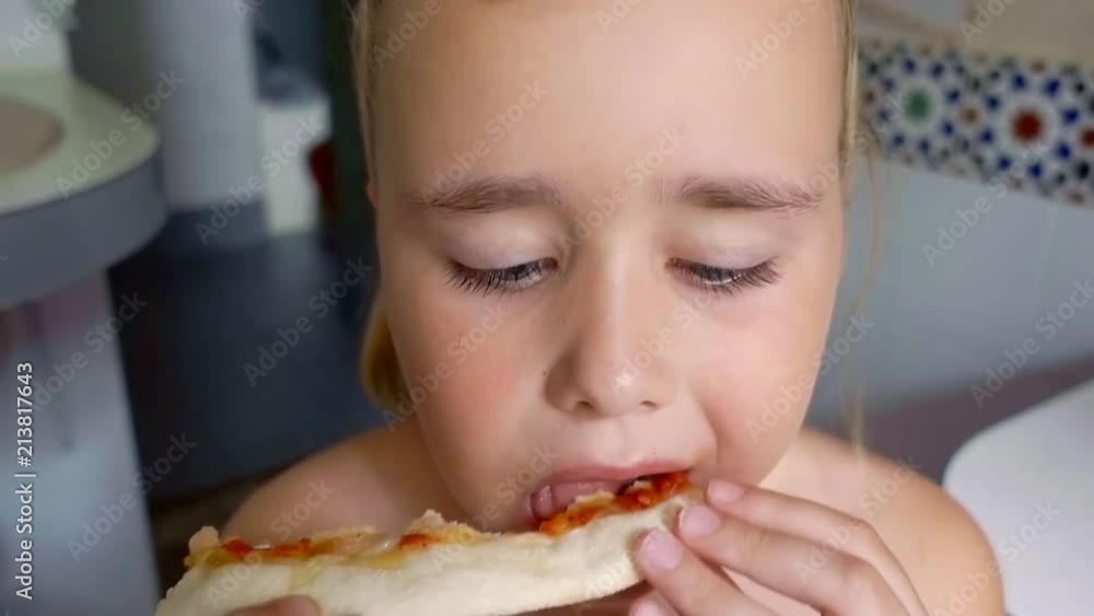 small hungry preschooler girl is biting and chewing piece of juicy tasty pizza in a room, close-up