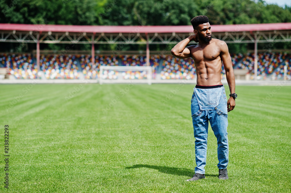 Handsome sexy african american muscular bare torso man at jeans overalls posed at green grass of stadium football field. Fashionable black man portrait.