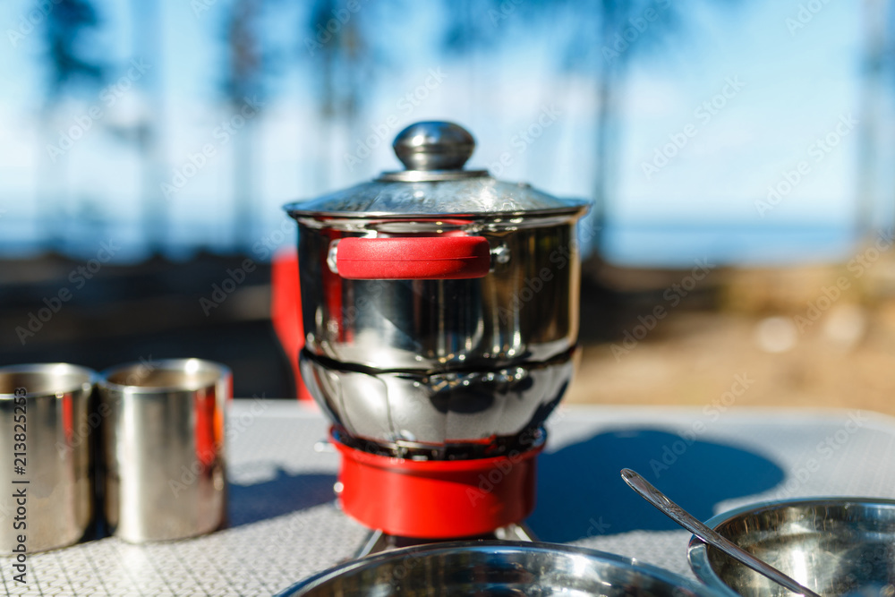 Aluminum pan boil on fire from a portable gas burner next to plates and mugs on a portable table against the background of the red folding chairs in the camping. Cooking while traveling by car. Aluminum pan boil on fire from a portable gas burner next to plates and mugs on a portable table against the background of the red folding chairs in the camping. Cooking while traveling by car.