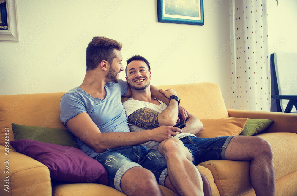 Two gay men in casual outfits sitting on sofa embracing and watching TV at home