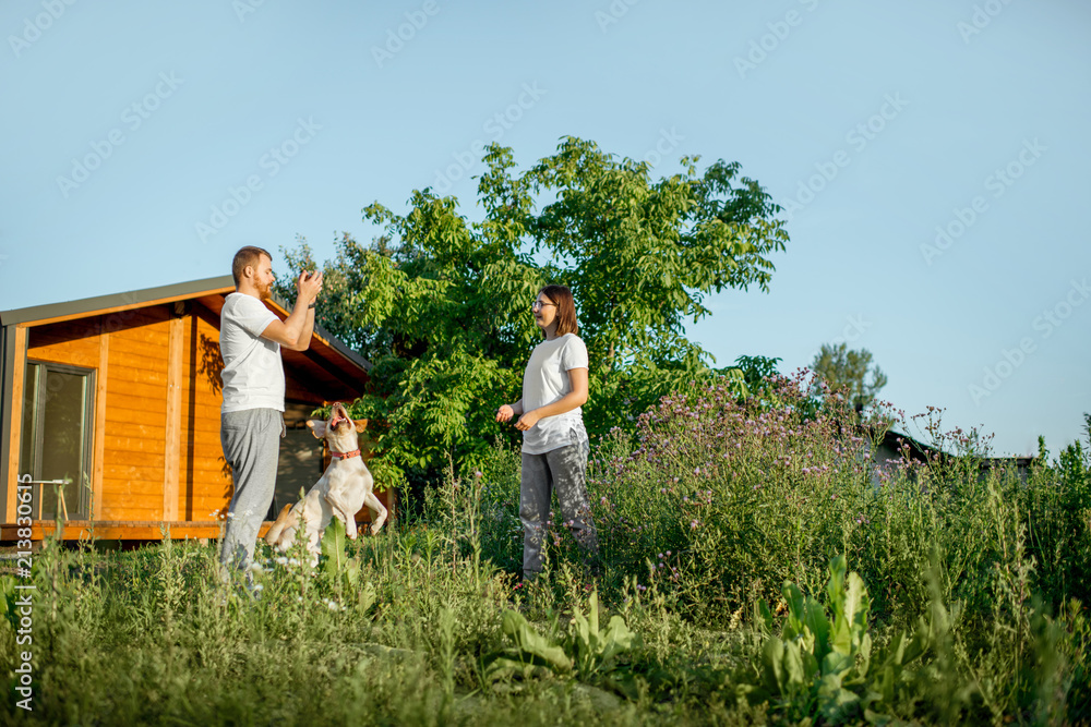 Young couple playing with dog on the backyard of the wooden country house during the sunset