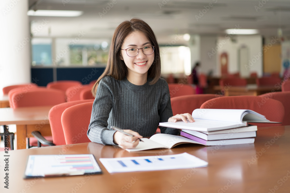Female student taking notes from a book at library, Young asian woman sitting at table doing assignments in college library Female student taking notes from a book at library, Young asian woman sitting at table doing assignments in college library