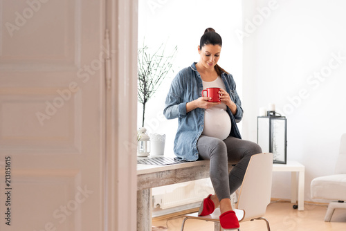 Принти на полотні A beautiful smiling pregnant woman sitting on a kitchen table in a denim jacket and drinking a coffee from a red mug
